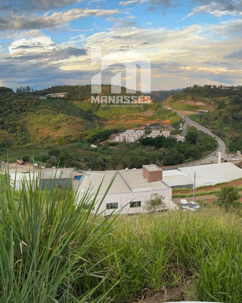 Vista panorâmica de um bairro em meio a morros e vegetação densa, com construções residenciais e industriais ao fundo, sob um céu parcialmente nublado ao entardecer. Logotipo da imobiliária Manassés sobreposto ao centro da imagem