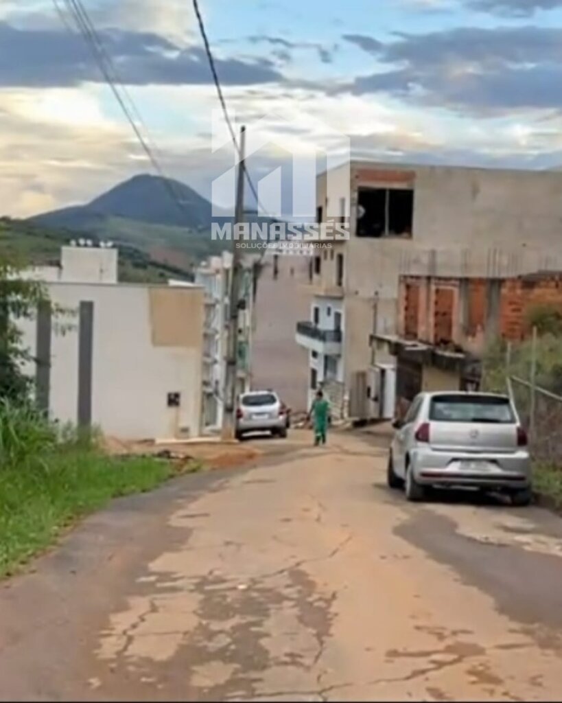 Rua de bairro em desenvolvimento com pavimento desgastado, dois carros estacionados, uma pessoa caminhando e construções residenciais em diferentes fases de acabamento. Ao fundo, montanha coberta por vegetação e céu parcialmente nublado. Logotipo "Manassés Soluções Imobiliárias" centralizado na imagem.