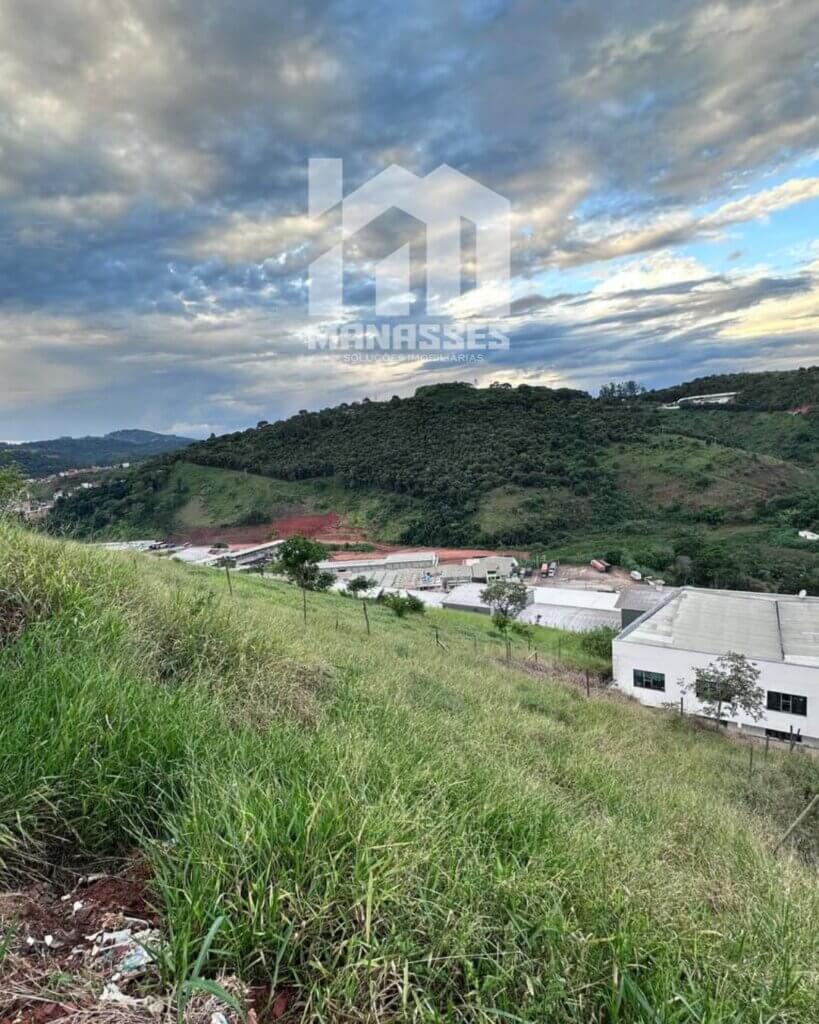 Terreno em declive coberto por vegetação rasteira, com vista para galpões industriais e colinas verdes ao fundo, sob um céu parcialmente nublado com tons azulados e cinzentos. Logotipo da Manassés Soluções Imobiliárias centralizado na imagem.