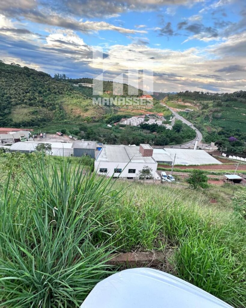 Vista de um terreno em declive coberto por vegetação alta, com uma pequena parte de um veículo visível no canto inferior. Ao fundo, galpões industriais, conjuntos residenciais e uma estrada sinuosa cercada por morros verdes sob um céu com nuvens e tons de azul. Logotipo da Manassés Soluções Imobiliárias sobreposto no centro da imagem.