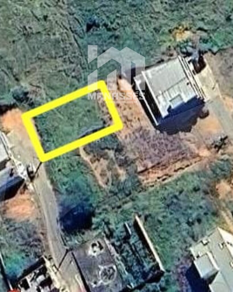 Imagem aérea de um terreno vazio demarcado por um contorno amarelo, localizado em um bairro com áreas verdes e construções ao redor. O lote está situado ao lado de uma rua pavimentada. Logotipo da Manassés Soluções Imobiliárias sobreposto no centro da imagem.