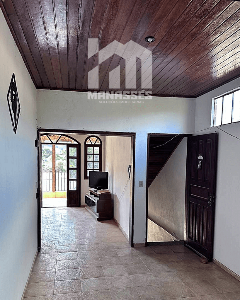 Sala de estar com piso cerâmico e teto de madeira, porta de entrada de madeira maciça, escada interna e acesso para varanda iluminada, em casa à venda pela Manassés Soluções Imobiliárias.