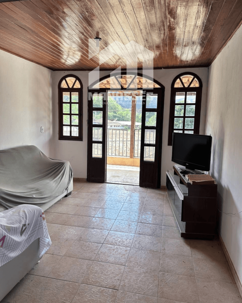 Sala iluminada com piso cerâmico, teto de madeira e portas de vidro com moldura de madeira, oferecendo acesso à varanda, em casa à venda pela Manassés Soluções Imobiliárias.