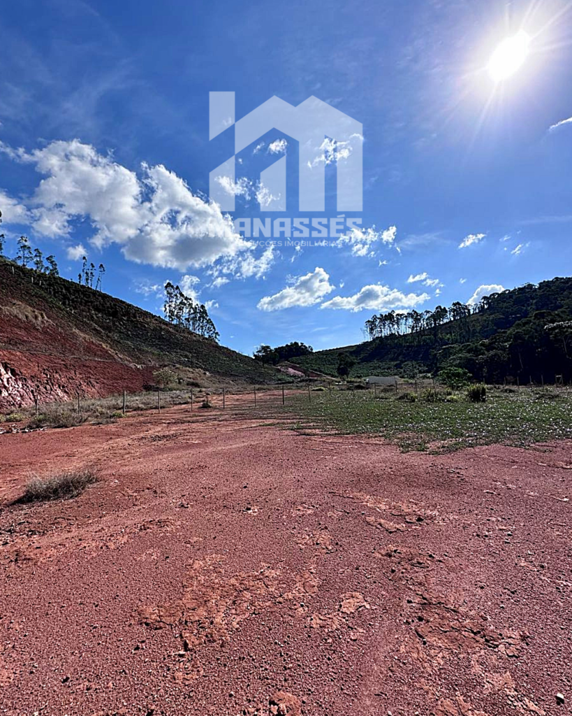 Terreno plano e ensolarado às margens da BR-262 em Manhuaçu/MG, com solo avermelhado, vista para morros verdes e céu azul aberto.